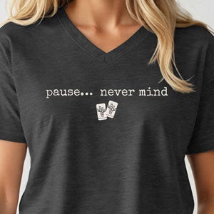 Pode incluir: Uma t-shirt cinzento-carv&atilde;o com decote em V, apresentando o texto branco "pause... never mind" impresso no peito, acompanhado por um pequeno gr&aacute;fico de duas pe&ccedil;as de mahjong brancas com um design de bambu centrado abaixo do texto.