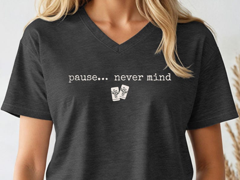 Pode incluir: Uma t-shirt cinzento-carv&atilde;o com decote em V, apresentando o texto branco "pause... never mind" impresso no peito, acompanhado por um pequeno gr&aacute;fico de duas pe&ccedil;as de mahjong brancas com um design de bambu centrado abaixo do texto.
