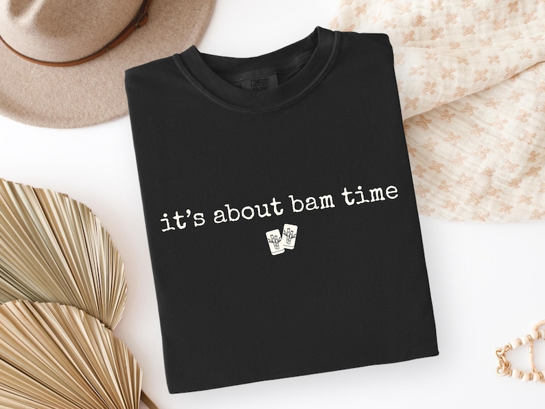 May include: Black crew neck t-shirt with the phrase "it's about bam time" in white text. The shirt also features a small graphic of two playing cards. The shirt is displayed on a white surface with a beige hat and decorative fan.