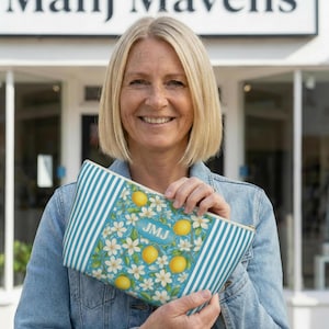 May include: A blue and white striped clutch with a lemon and floral pattern. The clutch has a rectangular shape and a zipper closure. The initials "JMJ" are visible on the front of the clutch. The background is a storefront.