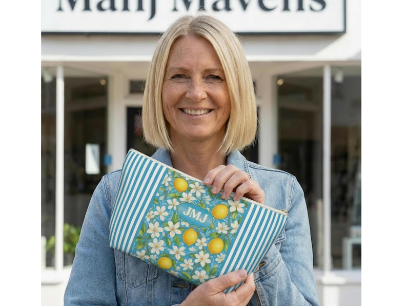 May include: A blue and white striped clutch with a lemon and floral pattern. The clutch has a rectangular shape and a zipper closure. The initials "JMJ" are visible on the front of the clutch. The background is a storefront.