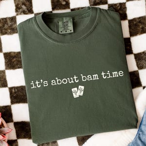 May include: Dark green t-shirt with the phrase "it's about bam time" printed in white. The shirt also features a small graphic of two drink glasses. The shirt is folded on a checkered surface with a pair of jeans visible.