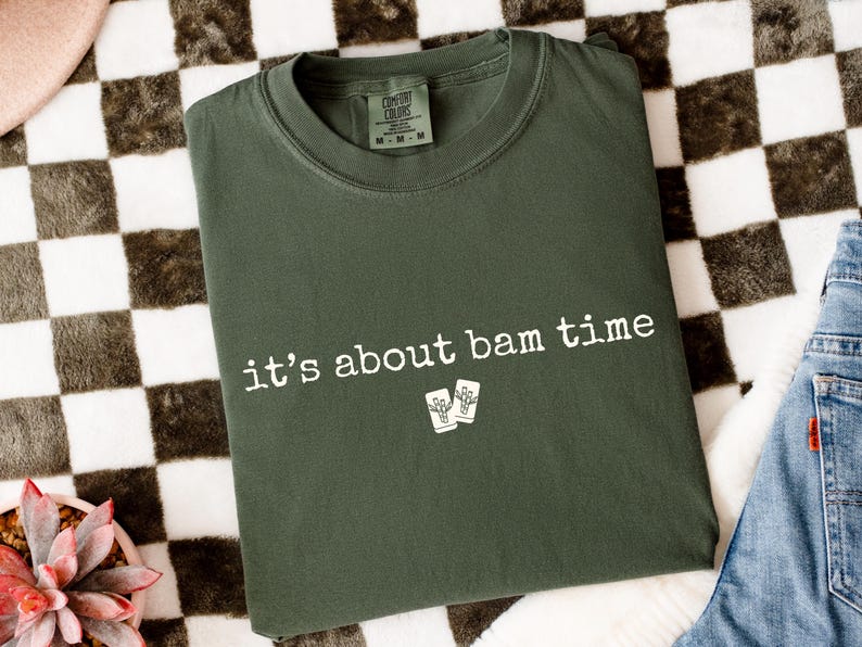 May include: Dark green t-shirt with the phrase "it's about bam time" printed in white. The shirt also features a small graphic of two drink glasses. The shirt is folded on a checkered surface with a pair of jeans visible.