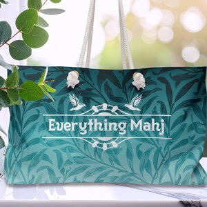 May include: A teal tote bag with a floral design and white rope handles. The bag displays the text "Everything Mahj" in a white banner, with bird and floral embellishments. The bag is placed on a white surface.