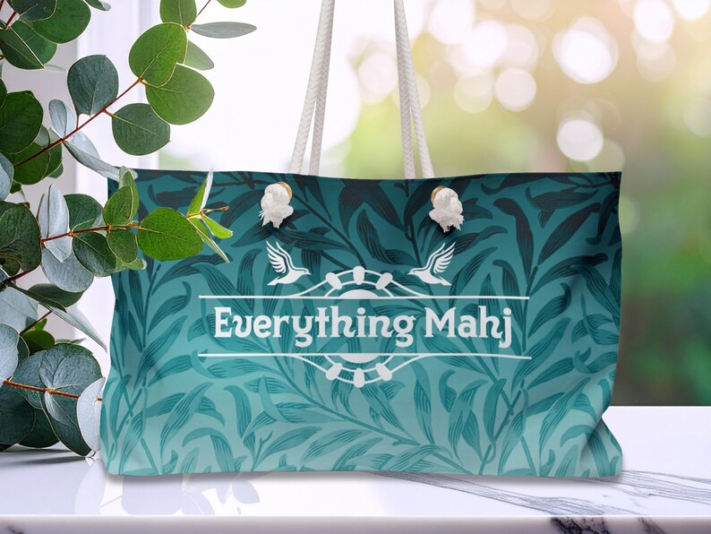 May include: A teal tote bag with a floral design and white rope handles. The bag displays the text "Everything Mahj" in a white banner, with bird and floral embellishments. The bag is placed on a white surface.