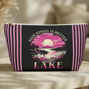 May include: Black zippered pouch with pink vertical stripes and a circular graphic. The graphic features a pink sunset over a lake with the text "MAH JONGG IS BETTER at the LAKE".