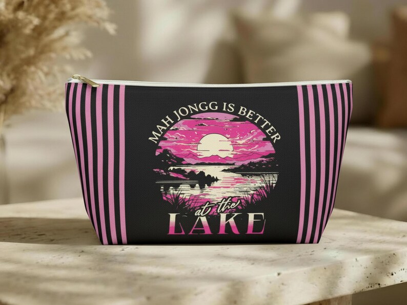 May include: Black zippered pouch with pink vertical stripes and a circular graphic. The graphic features a pink sunset over a lake with the text "MAH JONGG IS BETTER at the LAKE".