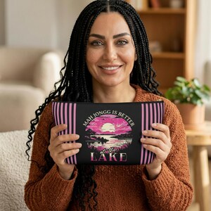 May include: Black zippered pouch with pink vertical stripes and a graphic of a sunset over a lake. The text on the pouch reads "Mah Jongg is Better at the Lake."