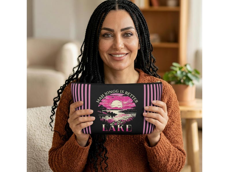 May include: Black zippered pouch with pink vertical stripes and a graphic of a sunset over a lake. The text on the pouch reads "Mah Jongg is Better at the Lake."