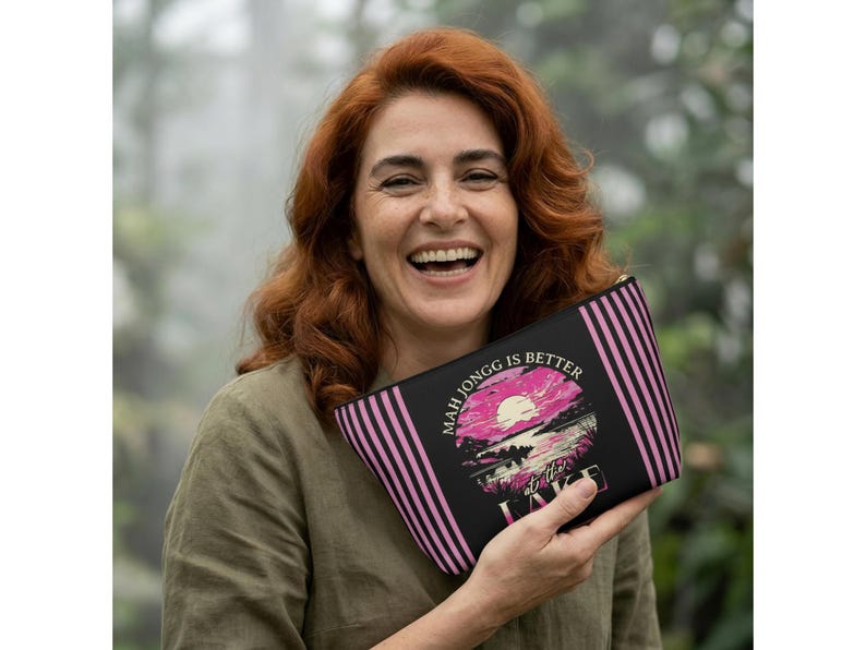 May include: Black zippered pouch with pink vertical stripes on the sides. The pouch features a graphic of a pink sun setting over a lake with the text "Mah Jongg is Better at the Lake."