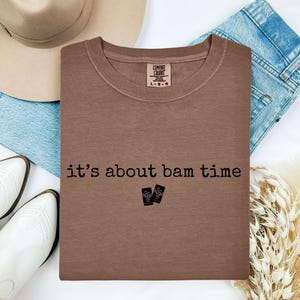 May include: A brown crew neck t-shirt with the phrase "it's about bam time" printed in black. The shirt is laid on top of blue jeans, a beige hat, and white boots. A tag on the shirt reads "Comfort Colors".