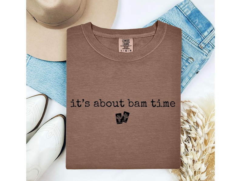 May include: A brown crew neck t-shirt with the phrase "it's about bam time" printed in black. The shirt is laid on top of blue jeans, a beige hat, and white boots. A tag on the shirt reads "Comfort Colors".