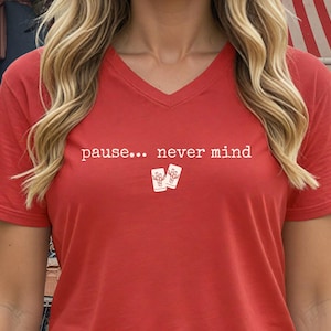 Pode incluir: Uma t-shirt vermelha com decote em V, apresentando o texto branco "pause... never mind" impresso no peito, acompanhado por um pequeno gr&aacute;fico de duas pe&ccedil;as retangulares brancas com um desenho linear de um bambu em cada pe&ccedil;a.