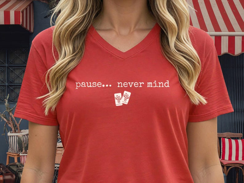 Pode incluir: Uma t-shirt vermelha com decote em V, apresentando o texto branco "pause... never mind" impresso no peito, acompanhado por um pequeno gr&aacute;fico de duas pe&ccedil;as retangulares brancas com um desenho linear de um bambu em cada pe&ccedil;a.
