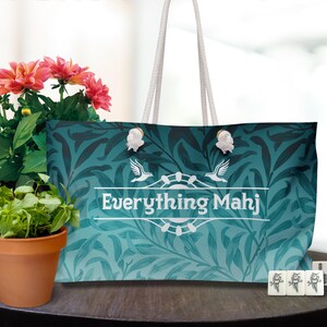 May include: A large tote bag with a teal and navy leaf pattern, the text "Everything Mahj", and white rope handles. The bag is next to a potted plant with pink flowers and a set of Mahjong tiles.