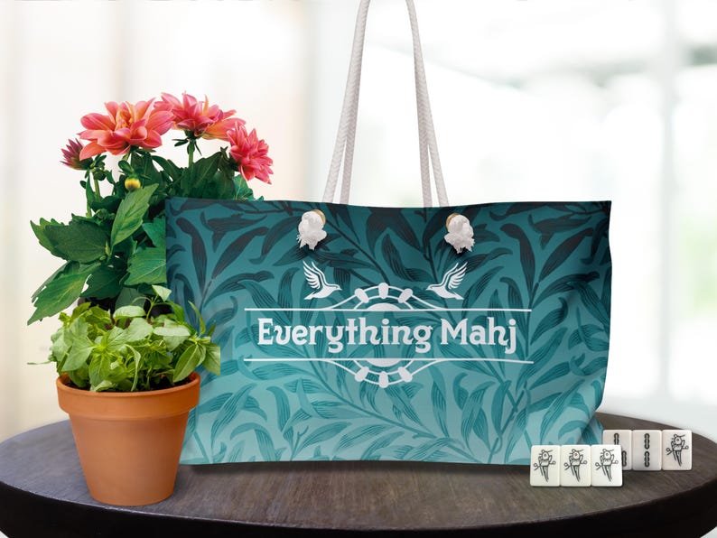 May include: A large tote bag with a teal and navy leaf pattern, the text "Everything Mahj", and white rope handles. The bag is next to a potted plant with pink flowers and a set of Mahjong tiles.