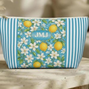 May include: A cosmetic bag with a blue and white striped pattern on the sides. The center features a design of lemons, white flowers, and the initials "JMJ" in a blue frame. The bag has a gold zipper.