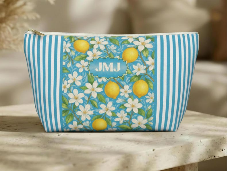 May include: A cosmetic bag with a blue and white striped pattern on the sides. The center features a design of lemons, white flowers, and the initials "JMJ" in a blue frame. The bag has a gold zipper.