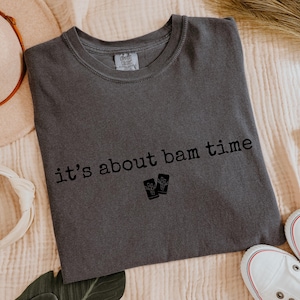 May include: Dark gray t-shirt with the text "it's about bam time" in black. The shirt features a crew neck and a small graphic of two objects. The shirt is laid flat on a textured surface, with a hat, headband, and shoes in the background.
