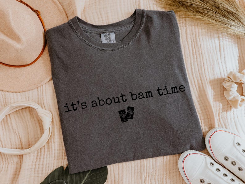 May include: Dark gray t-shirt with the text "it's about bam time" in black. The shirt features a crew neck and a small graphic of two objects. The shirt is laid flat on a textured surface, with a hat, headband, and shoes in the background.