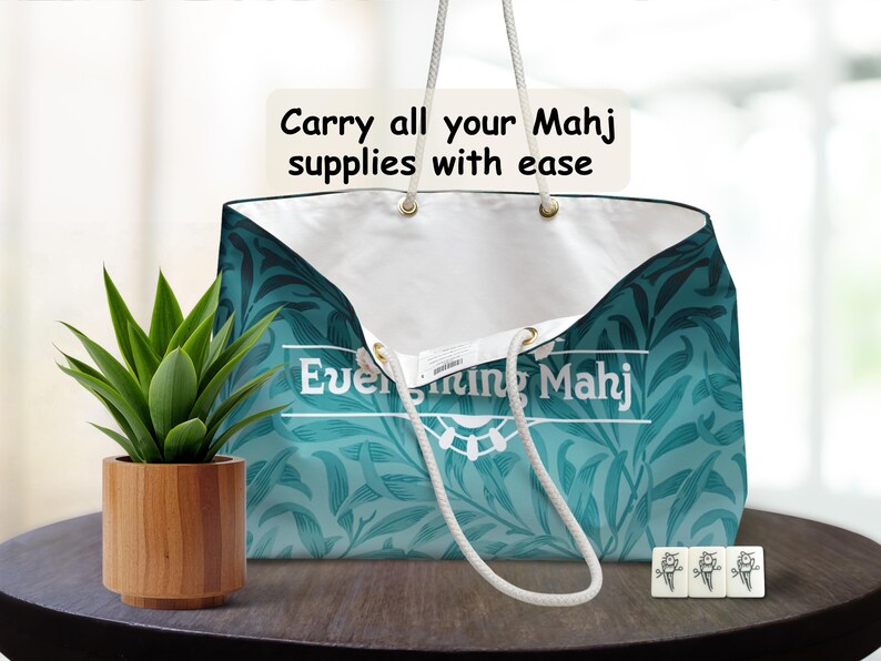 May include: A teal floral patterned tote bag with white interior, designed for Mahj supplies. It features white rope handles and text: "Carry all your Mahj supplies with ease" and "Everything Mahj". A small potted plant and Mahj tiles are on the table.