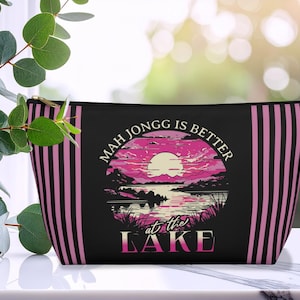 May include: Black zippered pouch with pink and black vertical stripes on the sides. The front features a circular graphic of a sunset over a lake with the text "Mah Jongg is Better at the Lake."