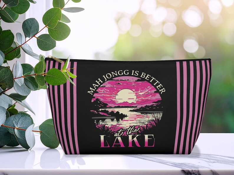 May include: Black zippered pouch with pink and black vertical stripes on the sides. The front features a circular graphic of a sunset over a lake with the text "Mah Jongg is Better at the Lake."