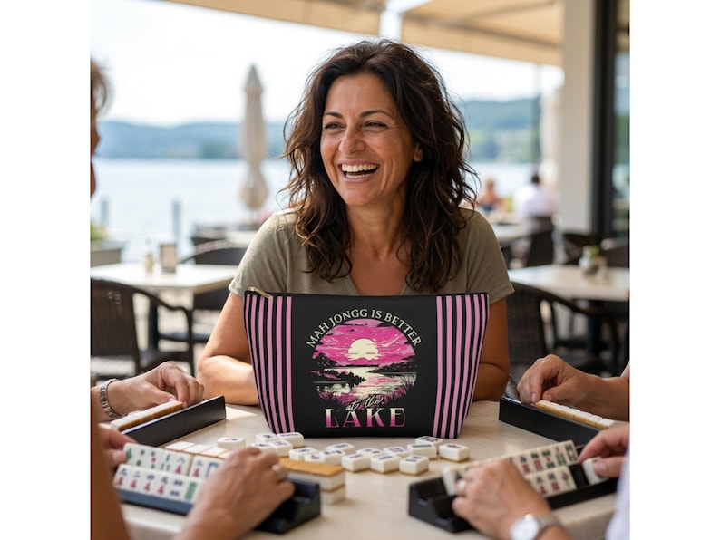 May include: A black and pink striped cosmetic bag with the text "Mah Jongg is Better at the Lake" and a sunset graphic. The bag is held by a person smiling while playing a game of Mahjongg.