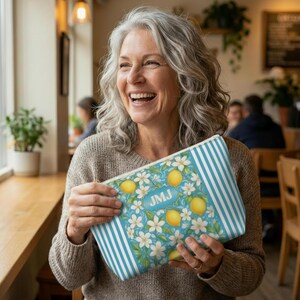 May include: A rectangular cosmetic bag with a blue and white striped pattern on the sides. The main design features lemons, white flowers, and the initials "JMJ" on a light blue background. The bag is held by a person.