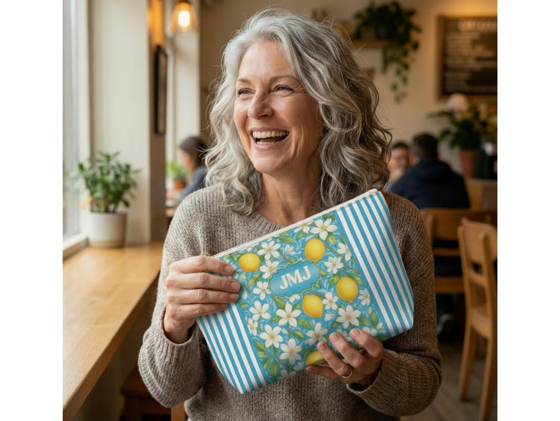May include: A rectangular cosmetic bag with a blue and white striped pattern on the sides. The main design features lemons, white flowers, and the initials "JMJ" on a light blue background. The bag is held by a person.