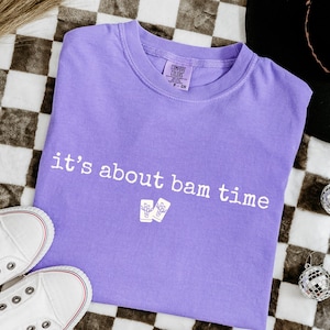 May include: A lavender Comfort Colors t-shirt with the phrase "it's about bam time" printed in white. The shirt is laid flat with white sneakers and disco ball ornaments nearby.