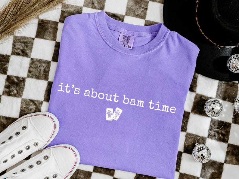 May include: A lavender Comfort Colors t-shirt with the phrase "it's about bam time" printed in white. The shirt is laid flat with white sneakers and disco ball ornaments nearby.