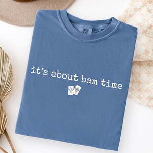 May include: A blue t-shirt with the phrase "it's about bam time" printed in white. The shirt also features a small graphic of two drinks. The shirt is folded and placed on a white surface.