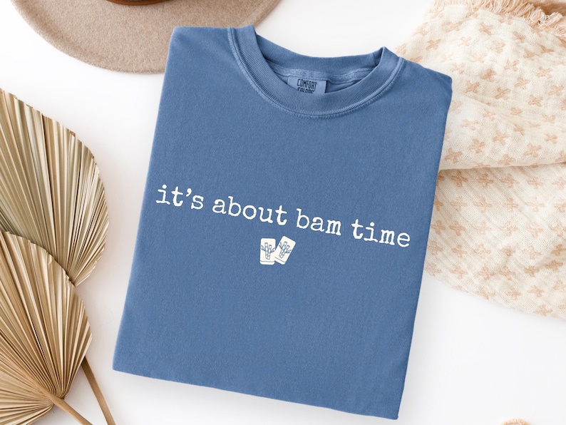 May include: A blue t-shirt with the phrase "it's about bam time" printed in white. The shirt also features a small graphic of two drinks. The shirt is folded and placed on a white surface.