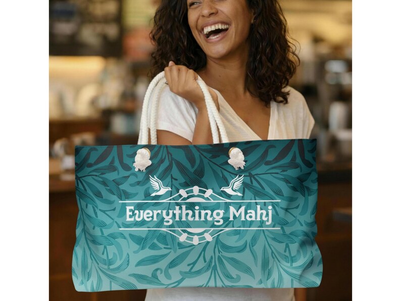 May include: A teal tote bag with a floral design and the text "Everything Mahj". The bag has white handles and is being held by a person. The bag's design features a gradient from teal to a lighter blue.