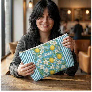 May include: A rectangular cosmetic bag with a blue and white striped pattern on the sides and a floral design with lemons and white flowers on a light blue background. The bag has a gold zipper and the initials "JMJ" in a blue rectangle.