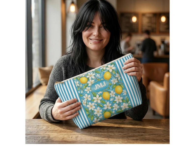 May include: A rectangular cosmetic bag with a blue and white striped pattern on the sides and a floral design with lemons and white flowers on a light blue background. The bag has a gold zipper and the initials "JMJ" in a blue rectangle.