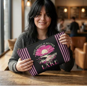 May include: A black zippered pouch with pink vertical stripes on the sides. The pouch features a graphic of a sunset over a lake with the text "MAH JONGG IS BETTER at the LAKE" in white and pink.
