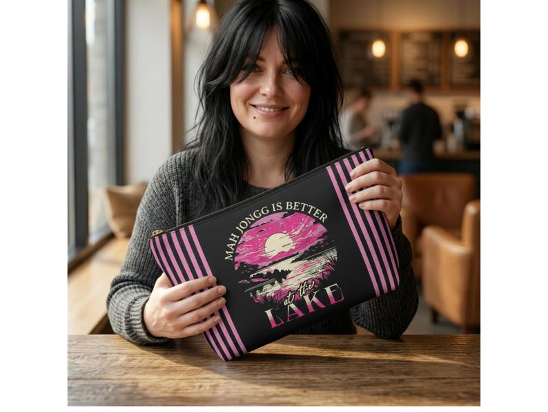 May include: A black zippered pouch with pink vertical stripes on the sides. The pouch features a graphic of a sunset over a lake with the text "MAH JONGG IS BETTER at the LAKE" in white and pink.