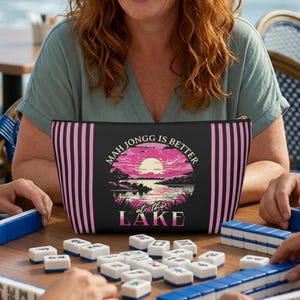 May include: Black zippered pouch with pink vertical stripes and a lake scene graphic. The text "MAH JONGG IS BETTER at THE LAKE" is displayed on the front. The pouch is on a wooden table with Mahjong tiles.