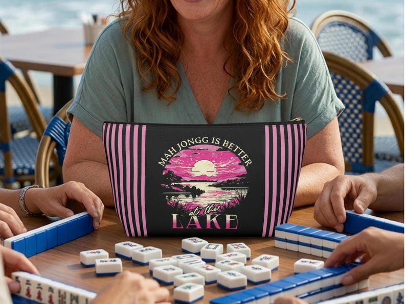 May include: Black zippered pouch with pink vertical stripes and a lake scene graphic. The text "MAH JONGG IS BETTER at THE LAKE" is displayed on the front. The pouch is on a wooden table with Mahjong tiles.