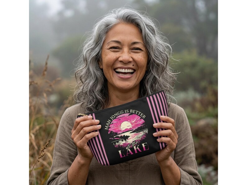 May include: Black zippered pouch with pink vertical stripes and the text "MAH JONGG IS BETTER at the LAKE" in white and pink. The pouch features a sunset scene with a pink sun, water, and trees.
