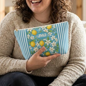 May include: A blue and white striped cosmetic bag with a lemon and floral design. The bag features a rectangular panel with lemons, white flowers, and the initials "JMJ" in a blue oval. The bag is held by a person wearing a beige sweater.