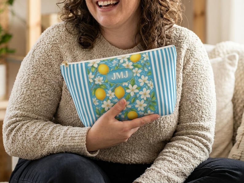 May include: A blue and white striped cosmetic bag with a lemon and floral design. The bag features a rectangular panel with lemons, white flowers, and the initials "JMJ" in a blue oval. The bag is held by a person wearing a beige sweater.