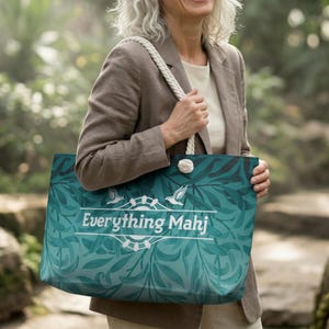 May include: A teal tote bag with a white rope handle and the text "Everything Mahj". The bag features a botanical design with a white emblem of a bird and a wheel. The bag is held by a person wearing a tan blazer.