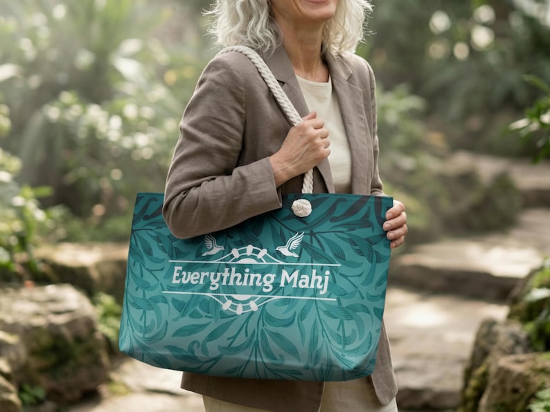 May include: A teal tote bag with a white rope handle and the text "Everything Mahj". The bag features a botanical design with a white emblem of a bird and a wheel. The bag is held by a person wearing a tan blazer.