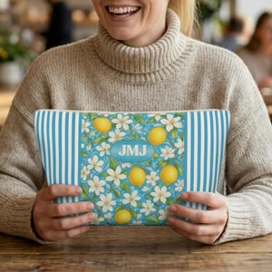 May include: A rectangular cosmetic bag with a blue and white striped pattern on the sides and a floral design in the center. The center design features lemons, white flowers, and the initials "JMJ" in a blue frame. The bag is held by a person.