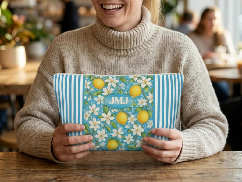 May include: A rectangular cosmetic bag with a blue and white striped pattern on the sides and a floral design in the center. The center design features lemons, white flowers, and the initials "JMJ" in a blue frame. The bag is held by a person.
