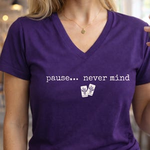 Pode incluir: Uma t-shirt roxa com decote em V apresentando o texto branco "pause... never mind" estampado no peito, acompanhado por um pequeno gr&aacute;fico de duas pe&ccedil;as de mahjong brancas com padr&otilde;es de bambu posicionadas abaixo do texto.