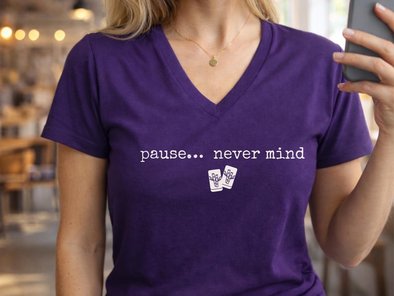Pode incluir: Uma t-shirt roxa com decote em V apresentando o texto branco "pause... never mind" estampado no peito, acompanhado por um pequeno gr&aacute;fico de duas pe&ccedil;as de mahjong brancas com padr&otilde;es de bambu posicionadas abaixo do texto.